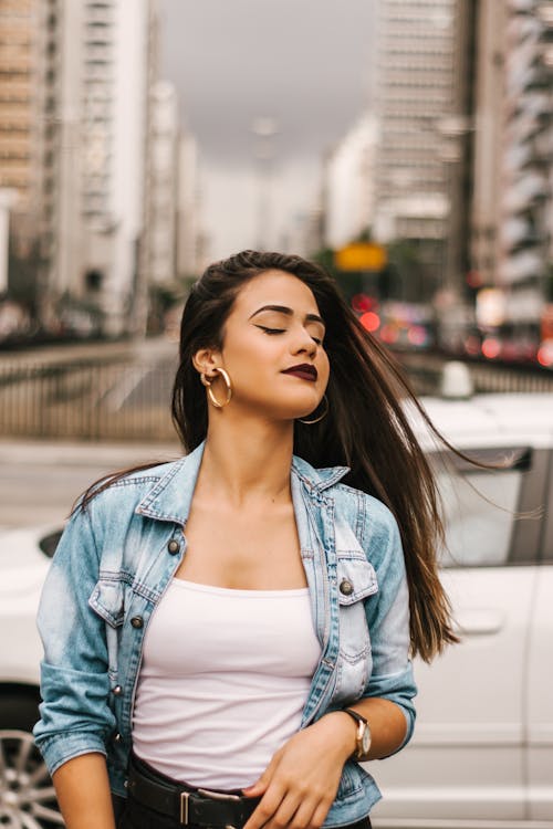Free Selective Focus Photo of Woman in Blue Denim Jacket, White Top, and Black Bottoms Posing In Front of White Car With Her Eyes Closed Stock Photo