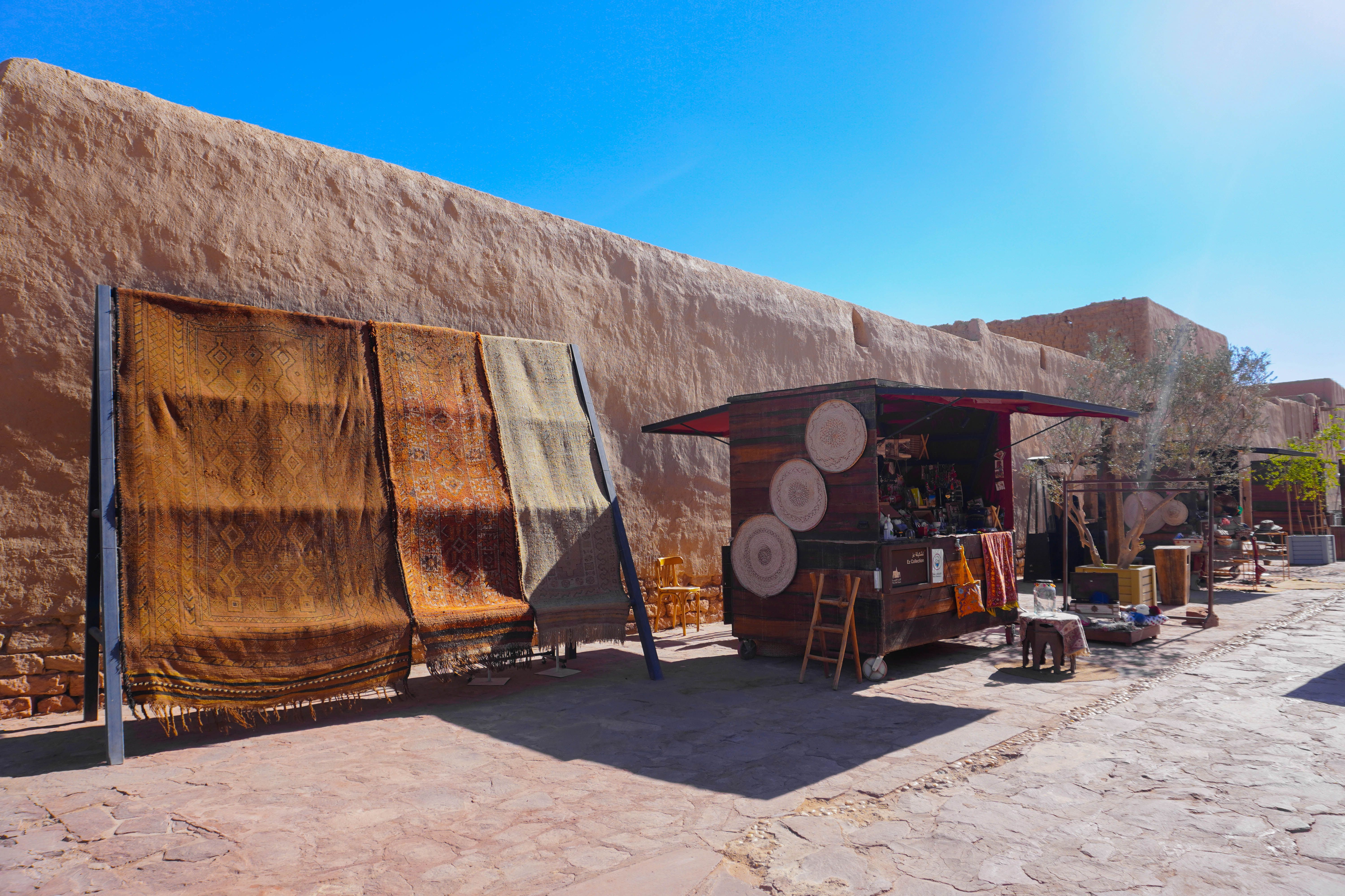 Market stall with woven rugs and baskets beside a long mud-brick wall