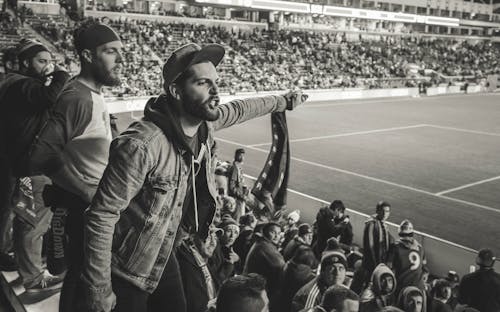 Монохромное фото человека в куртке, указывающего пальцем на стадионе