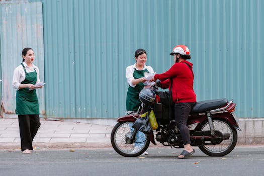 Two women in aprons hand out flyers to a motorcyclist on an urban street.