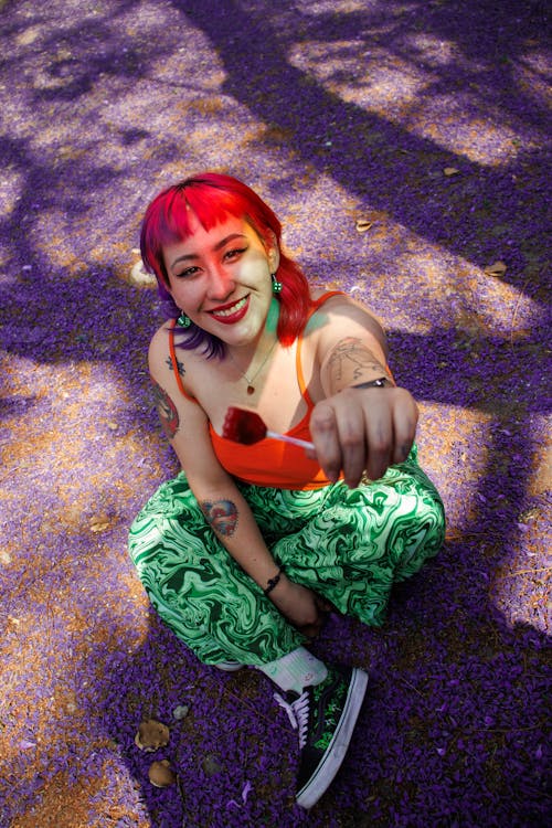 A woman with red hair and green hair is sitting on the ground