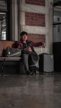 Senior man sits in a railway station waiting room with luggage, reflecting travel patience.