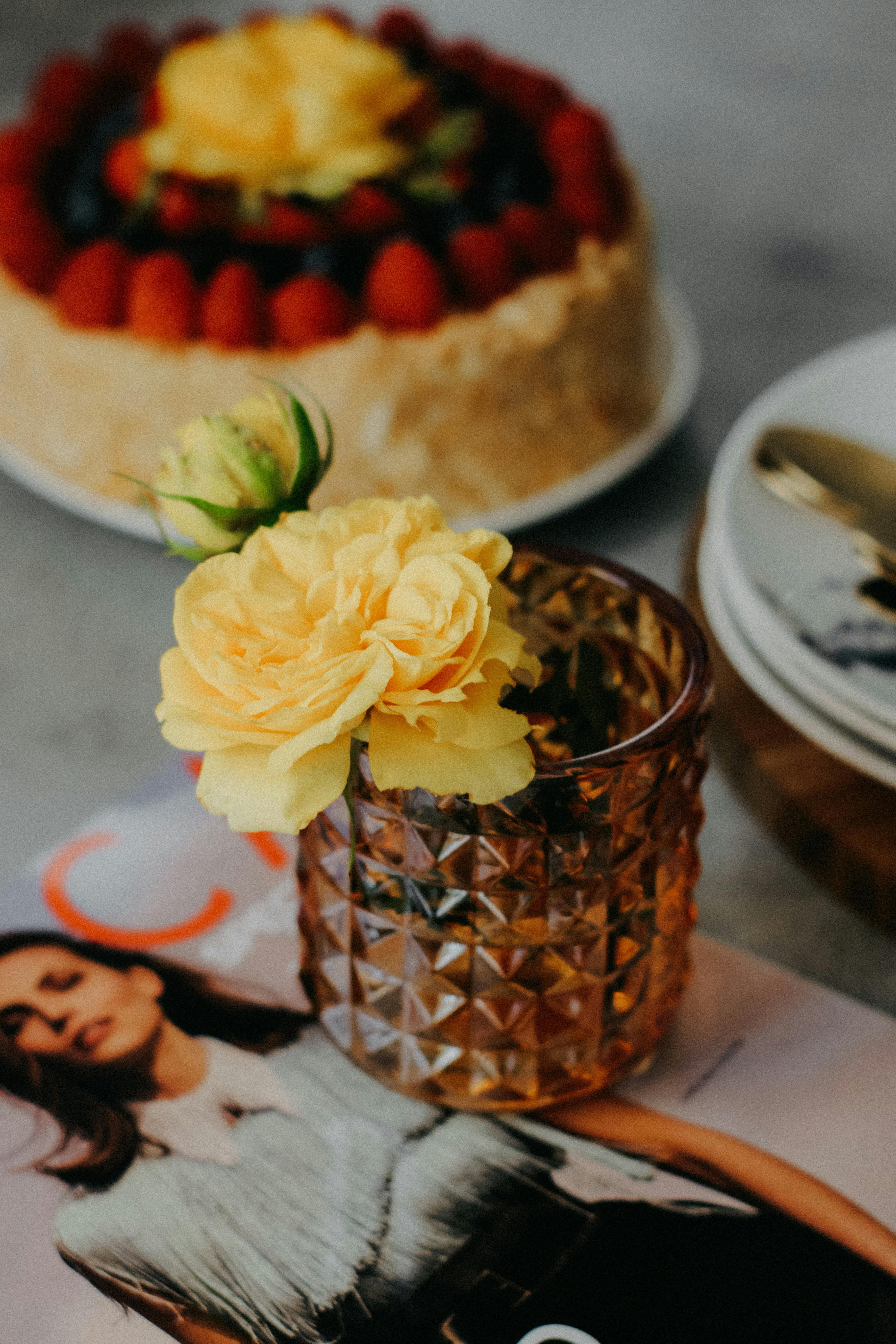 A stylish display of yellow roses in a glass vase and a fruit cake placed elegantly on a magazine.