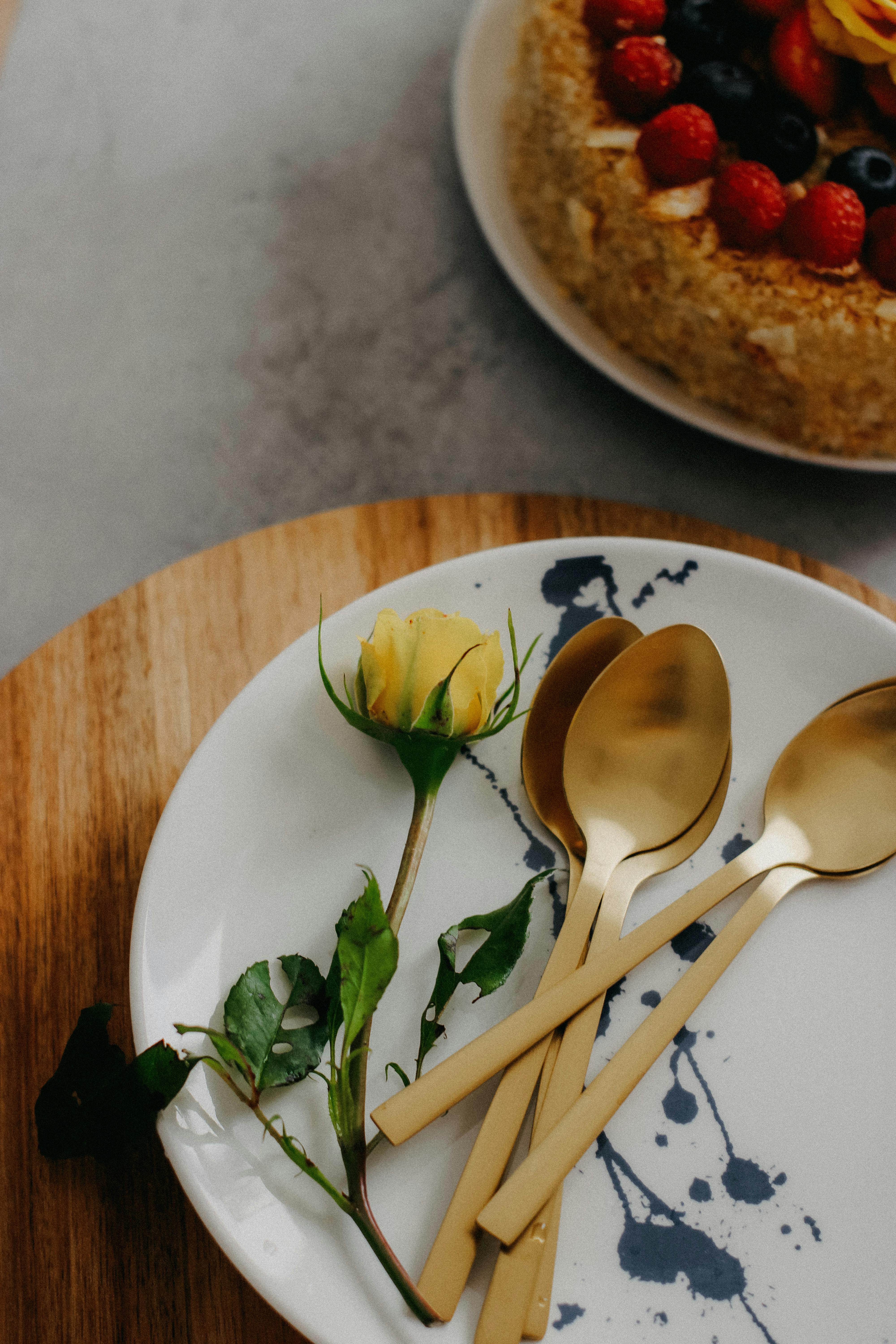 A delicious cake with berries next to golden spoons and a yellow rose on a decorated plate.