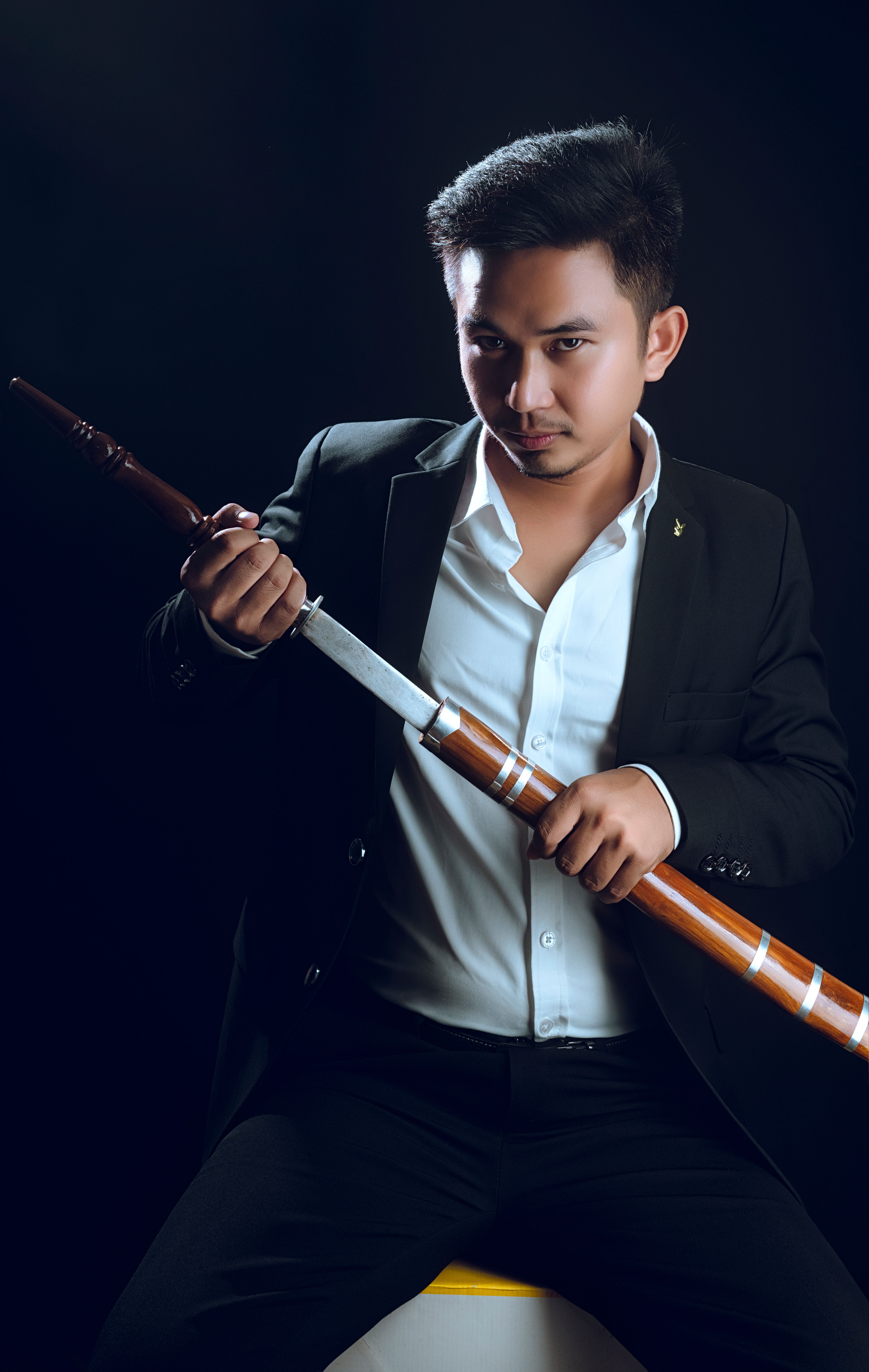 Stylish man in a black suit holding a sword against a dark background.
