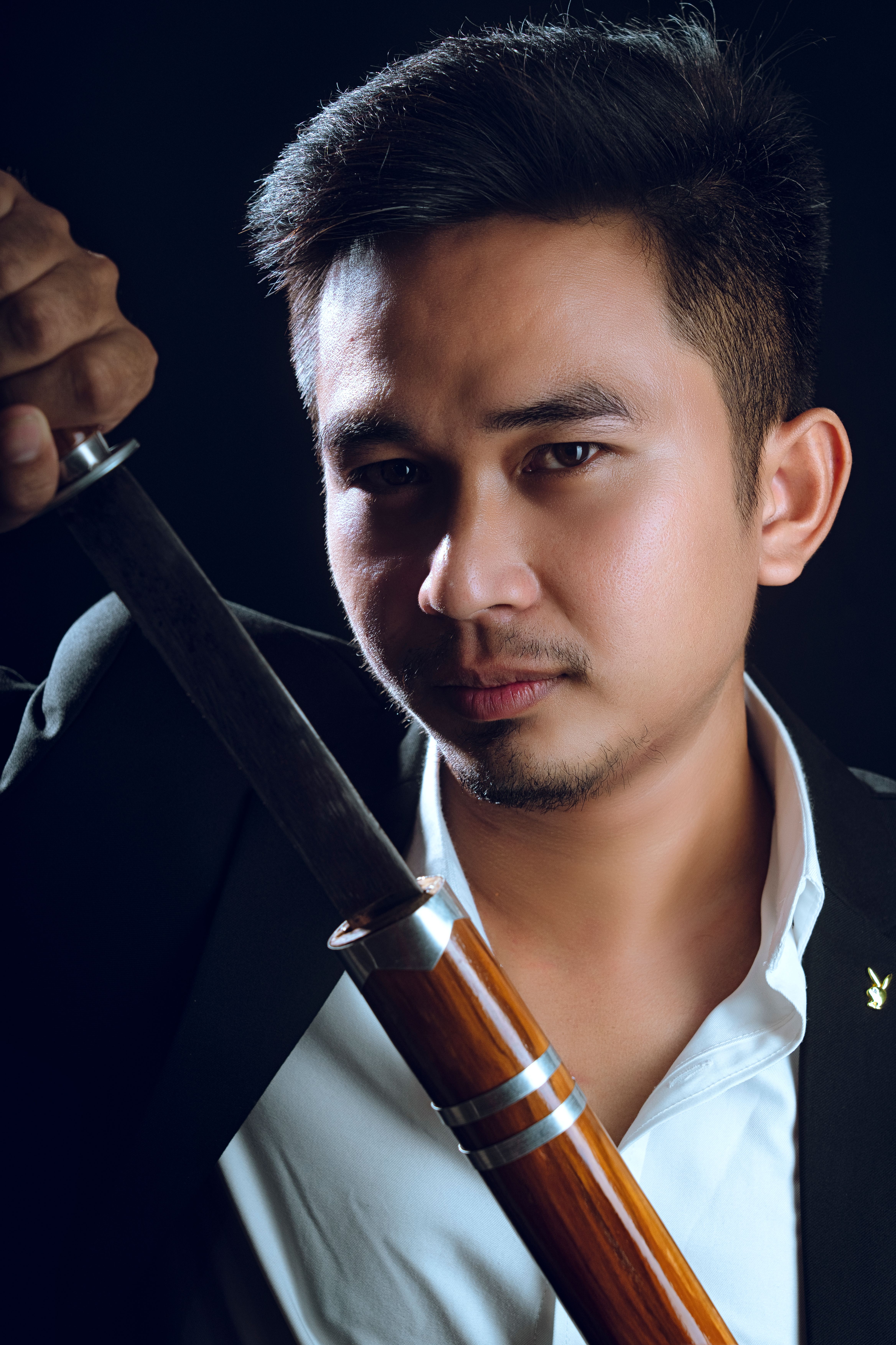 Asian man dressed elegantly holding a sword, posing confidently in a studio setting.
