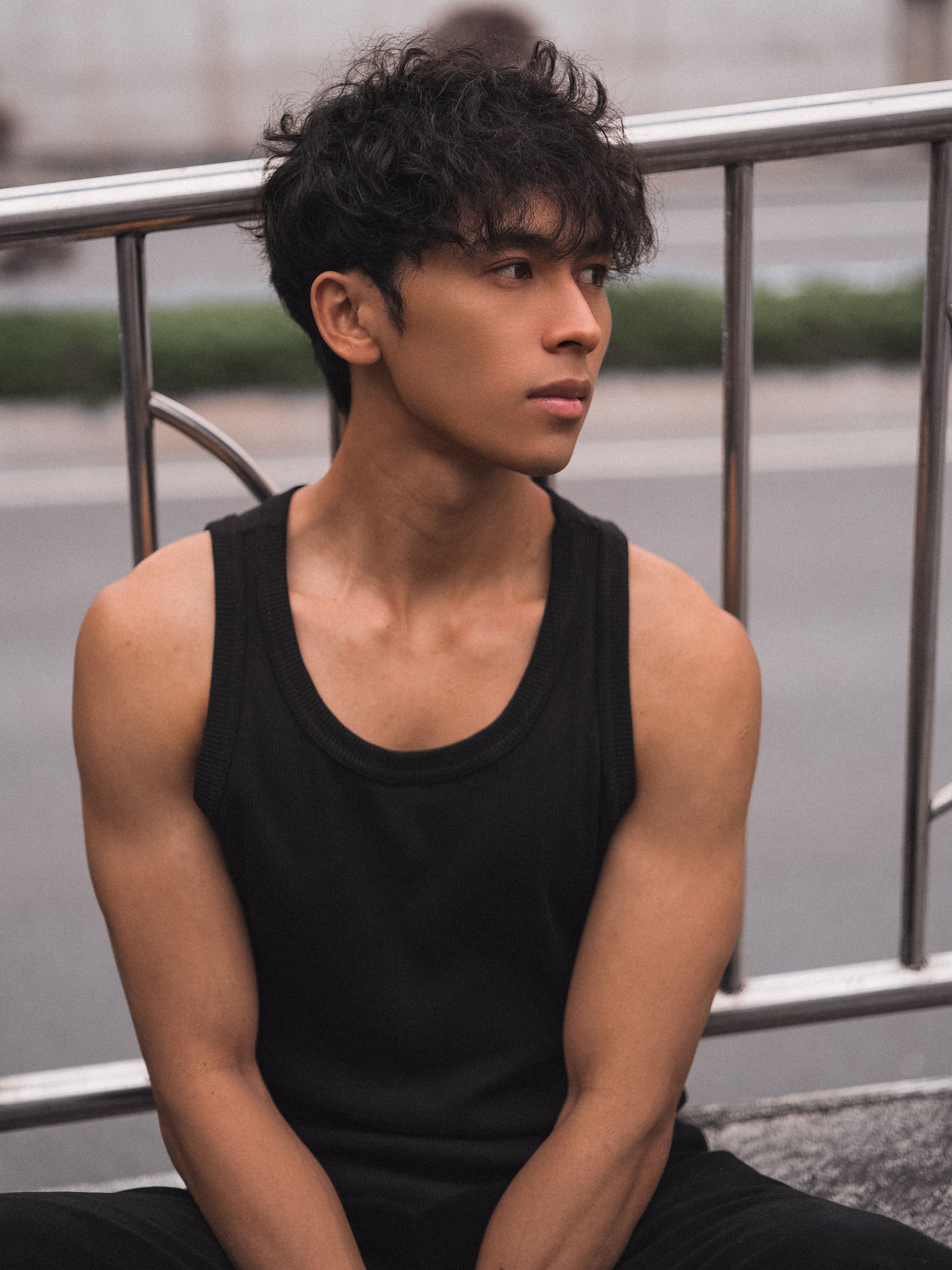 Stylish portrait of a young man in black attire sitting by a railing in an urban setting.