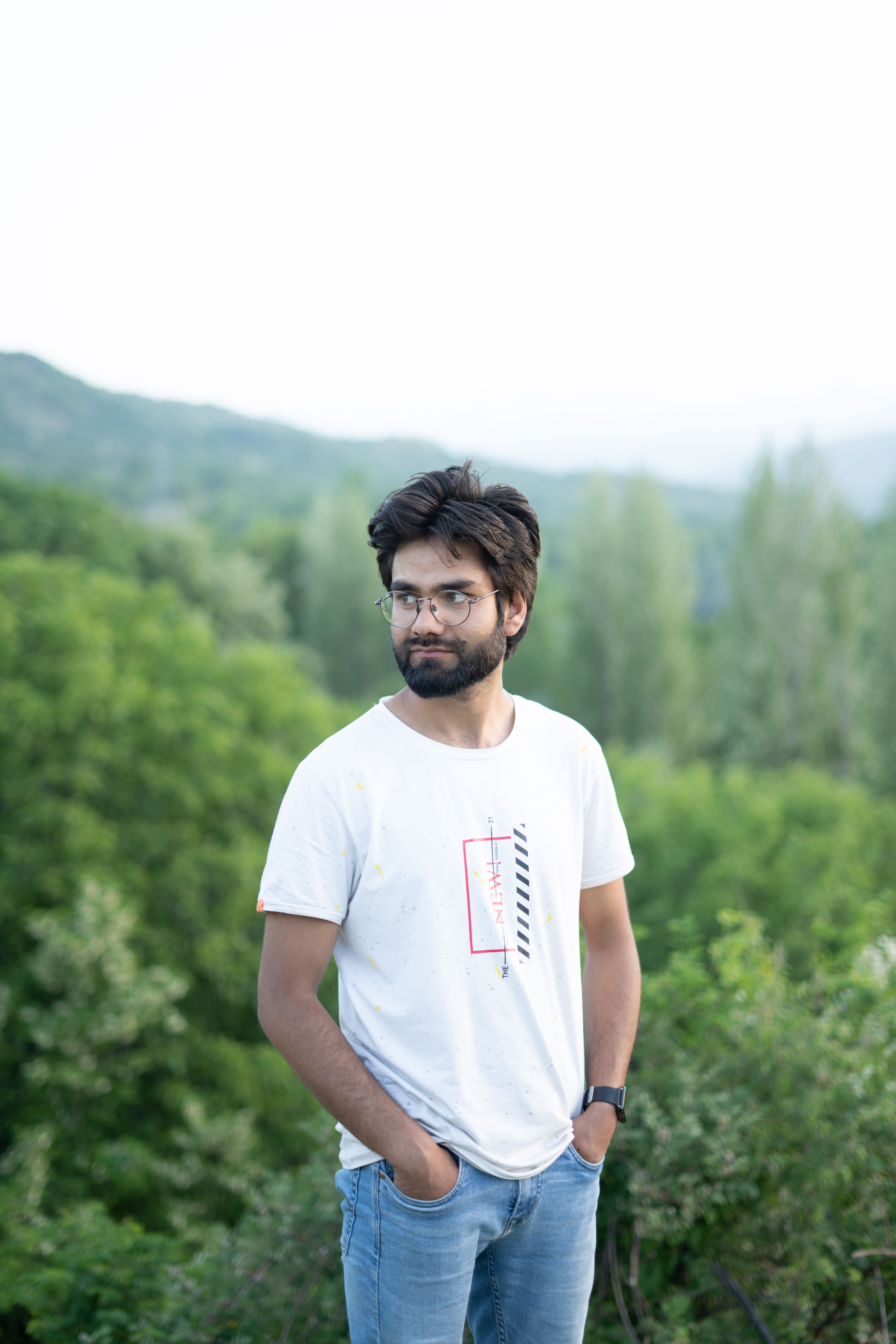 Stylish man with a beard and eyeglasses poses in a lush green landscape in Srinagar.