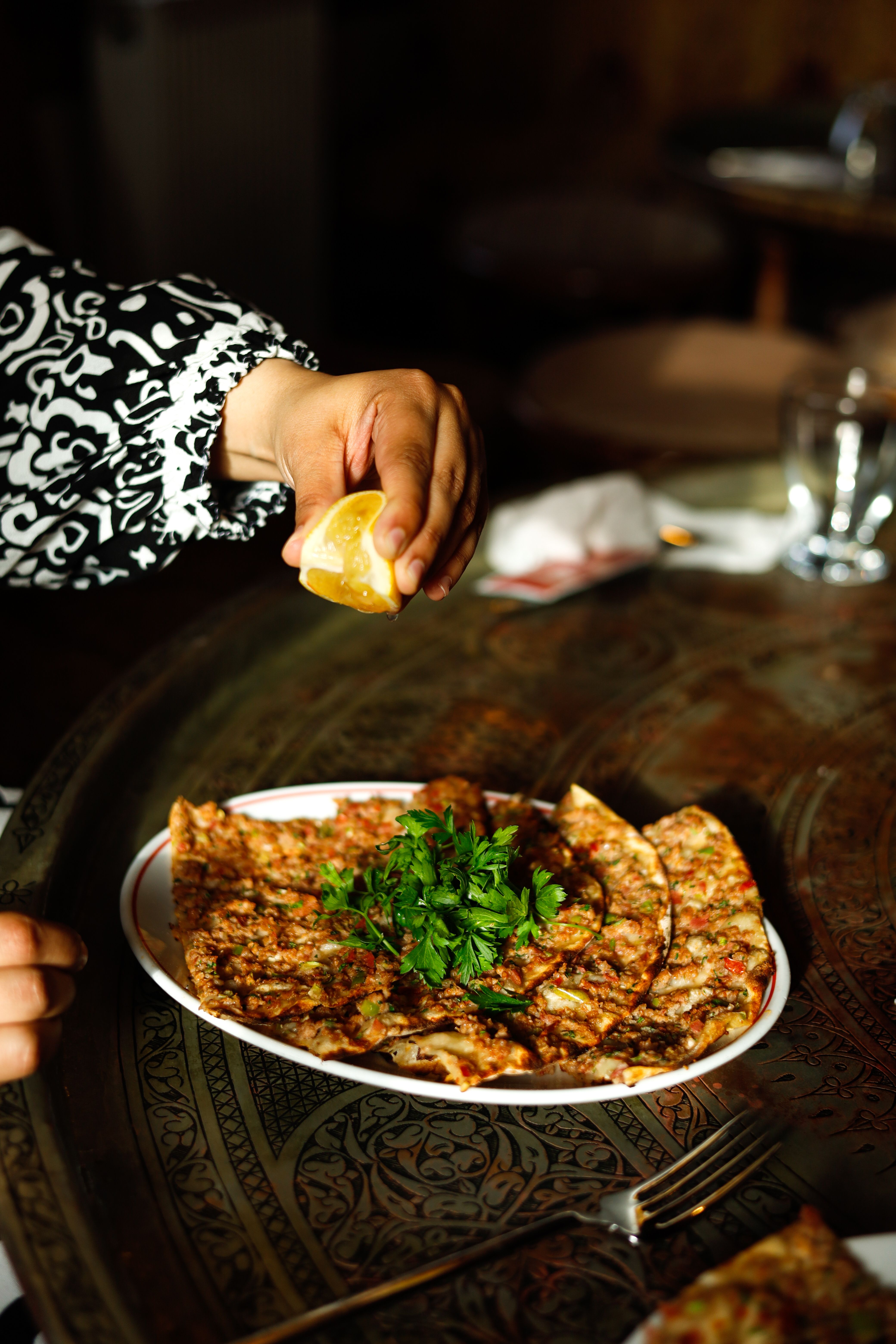 Main pressant un quartier de citron au-dessus d’une assiette de lahmacun au persil, posée sur un plateau de cuivre gravé