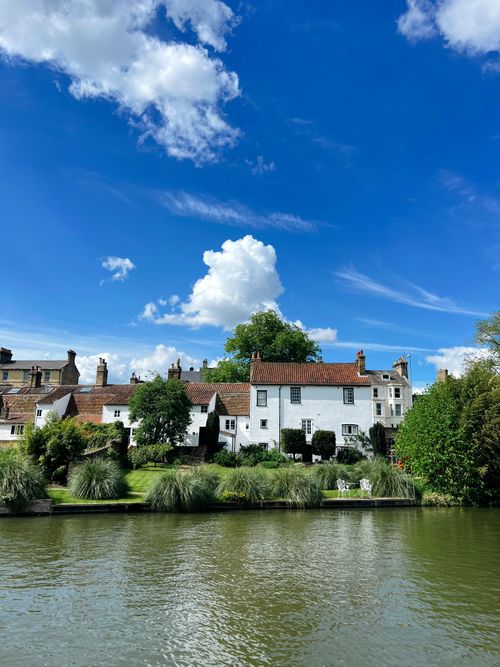 Free Scenic view of charming riverside cottages with lush greenery and blue sky. Stock Photo