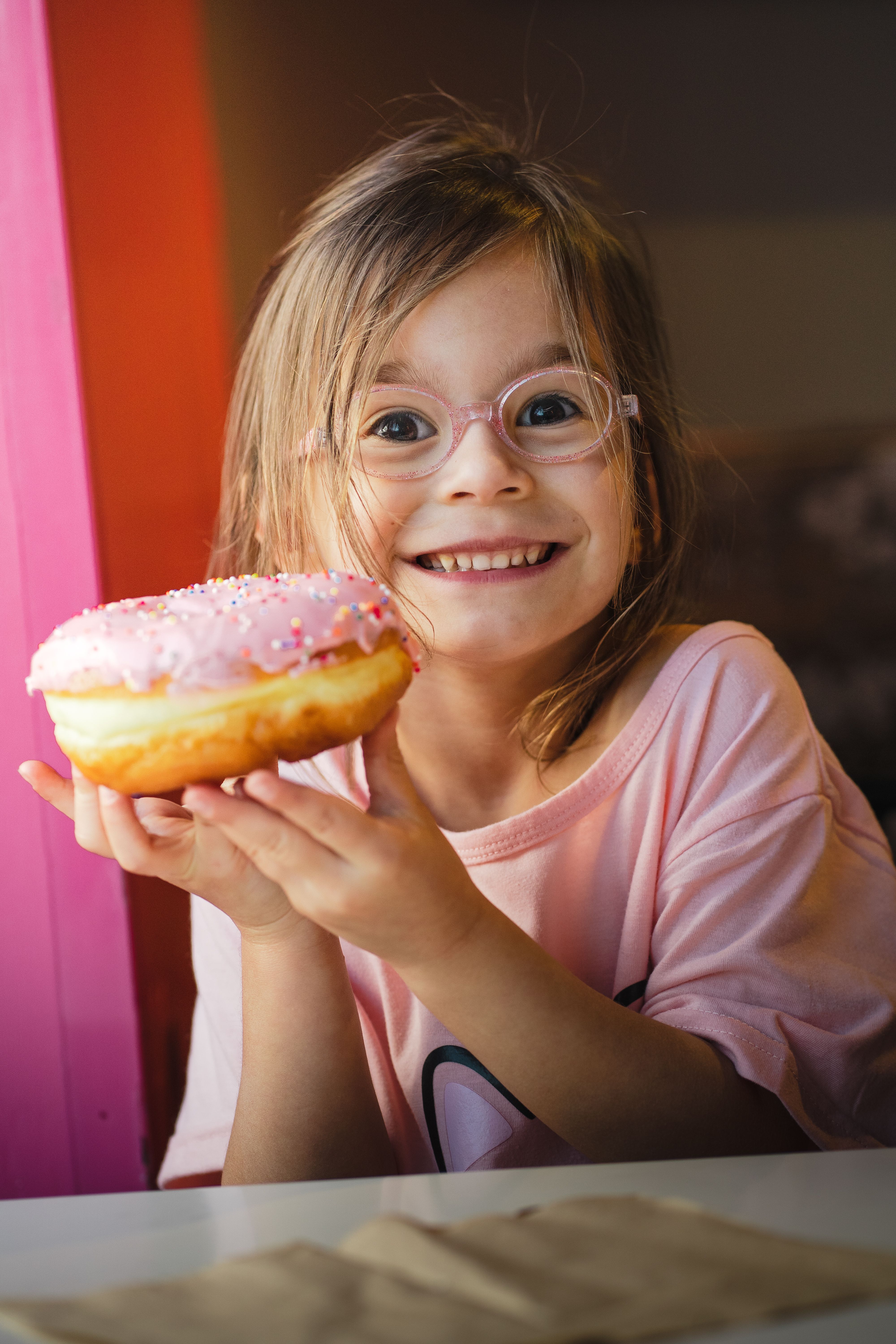 Smiling girl holding a delicious pink sprinkle donut captures pure delight in a warm indoor setting.