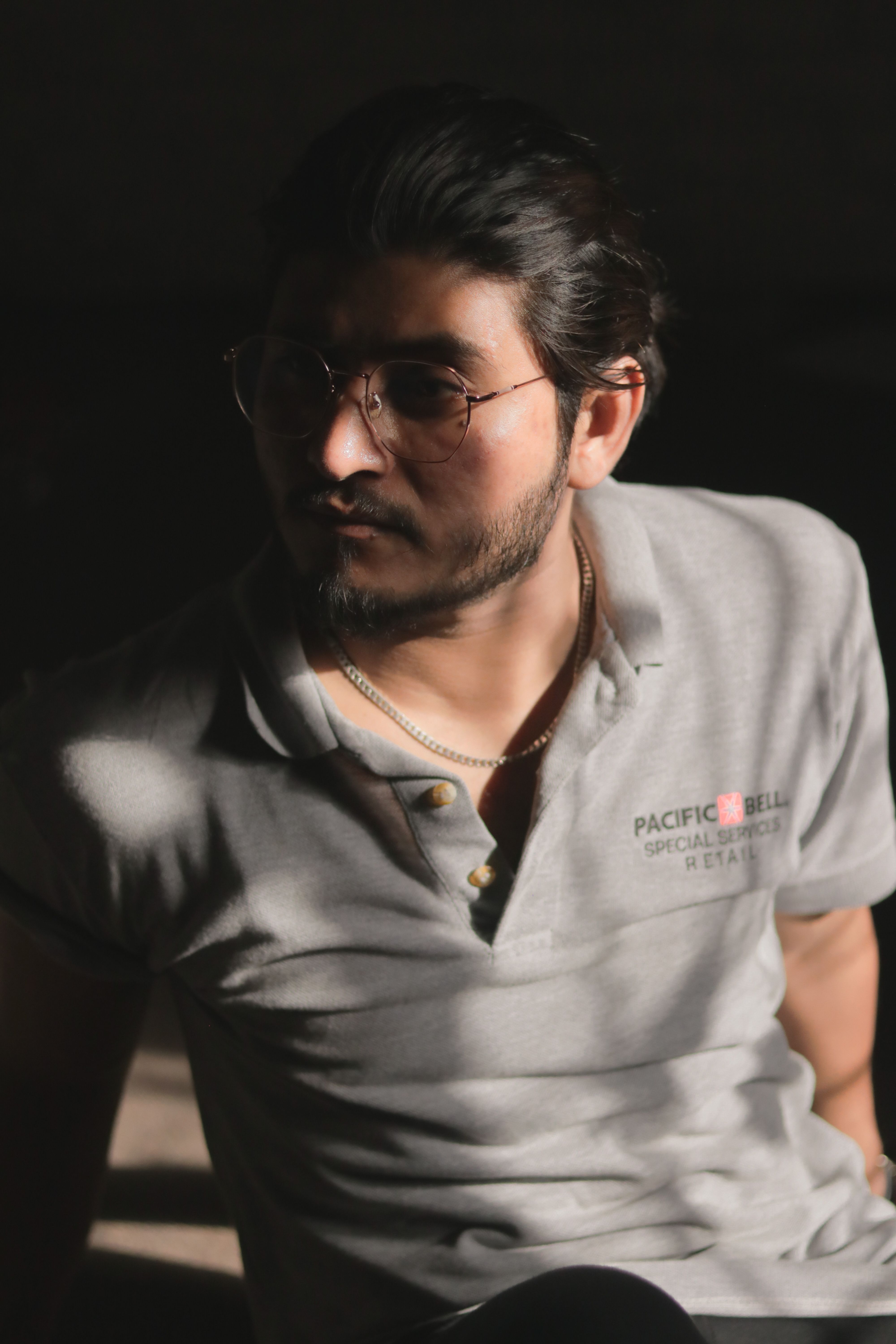 Backlit portrait of a young man wearing sunglasses and a grey polo shirt.