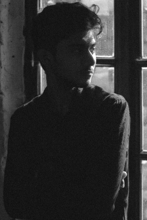 Free Black and white portrait of a young man standing by a window, creating a moody atmosphere. Stock Photo