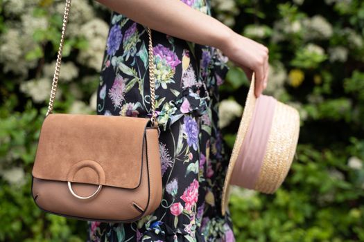 Stylish woman in floral dress holding a handbag and straw hat in a garden.