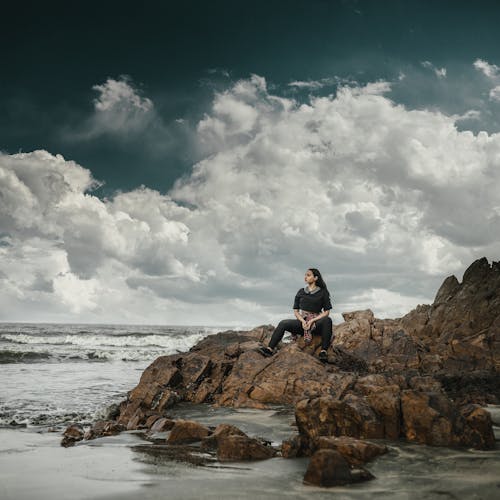 Foto Der Frau, Die Auf Felsen Nahe Strand Sitzt Und In Die Ferne Schaut