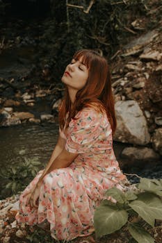 这幅宁静的肖像描绘了一位身着花卉连衣裙的女子，她正在河畔的自然风光中放松身心