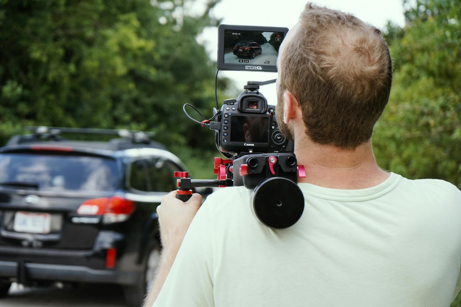 Videographer using shoulder-mounted camera rig to film vehicle scene outdoors in daylight.