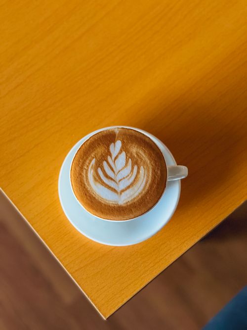 Free A cappuccino with intricate froth art served on a white plate atop a wooden table viewed from above. Stock Photo