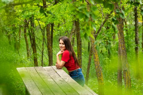 Free Smiling young woman in red shirt posing on a wooden bench surrounded by lush green forest. Stock Photo