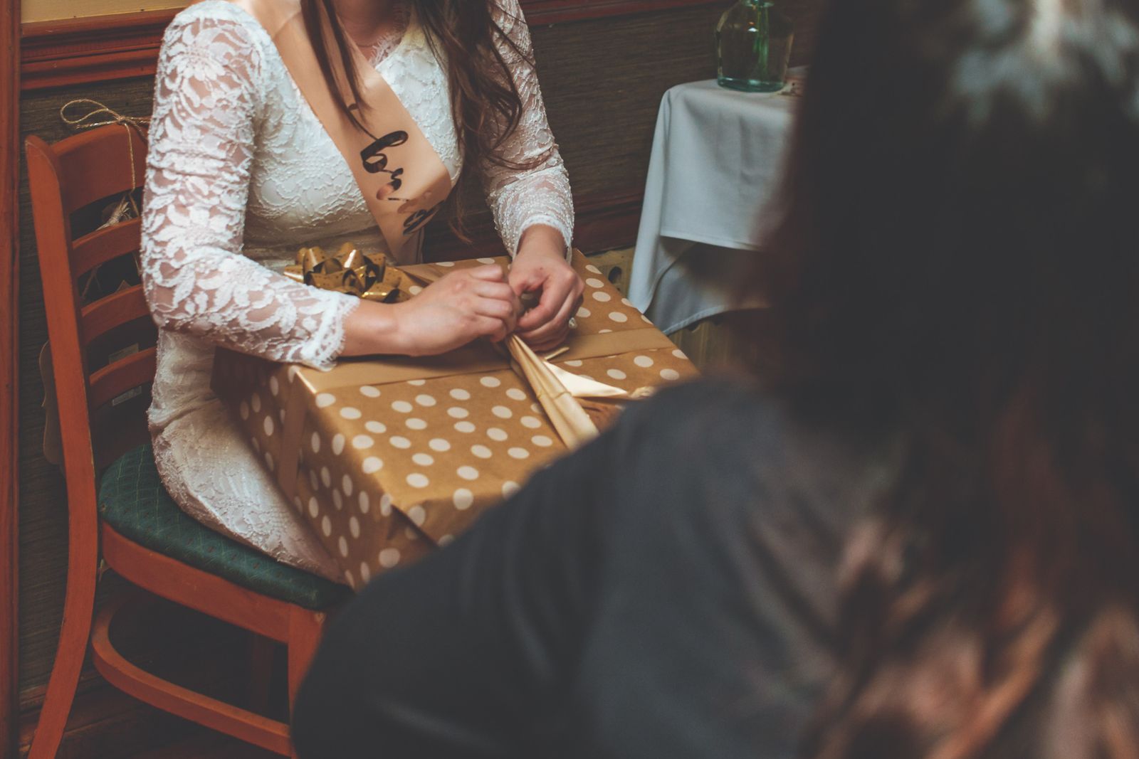 A woman at a bridal shower unwrapping a polka-dot gift indoors while wearing a bride-to-be sash