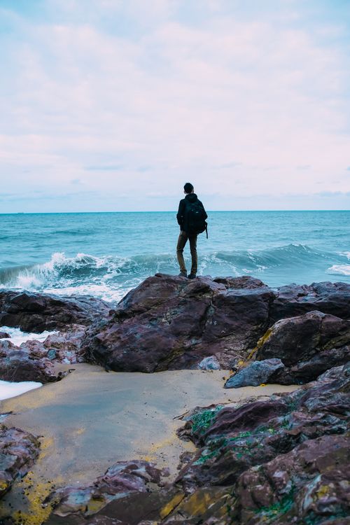 Free A lone traveler with a backpack stands on a rocky coastline, facing the ocean waves. Stock Photo