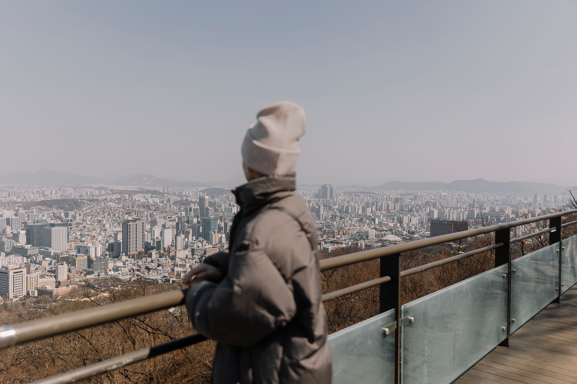 A woman in warm clothing overlooks the Seoul cityscape from an observation deck on a clear day.