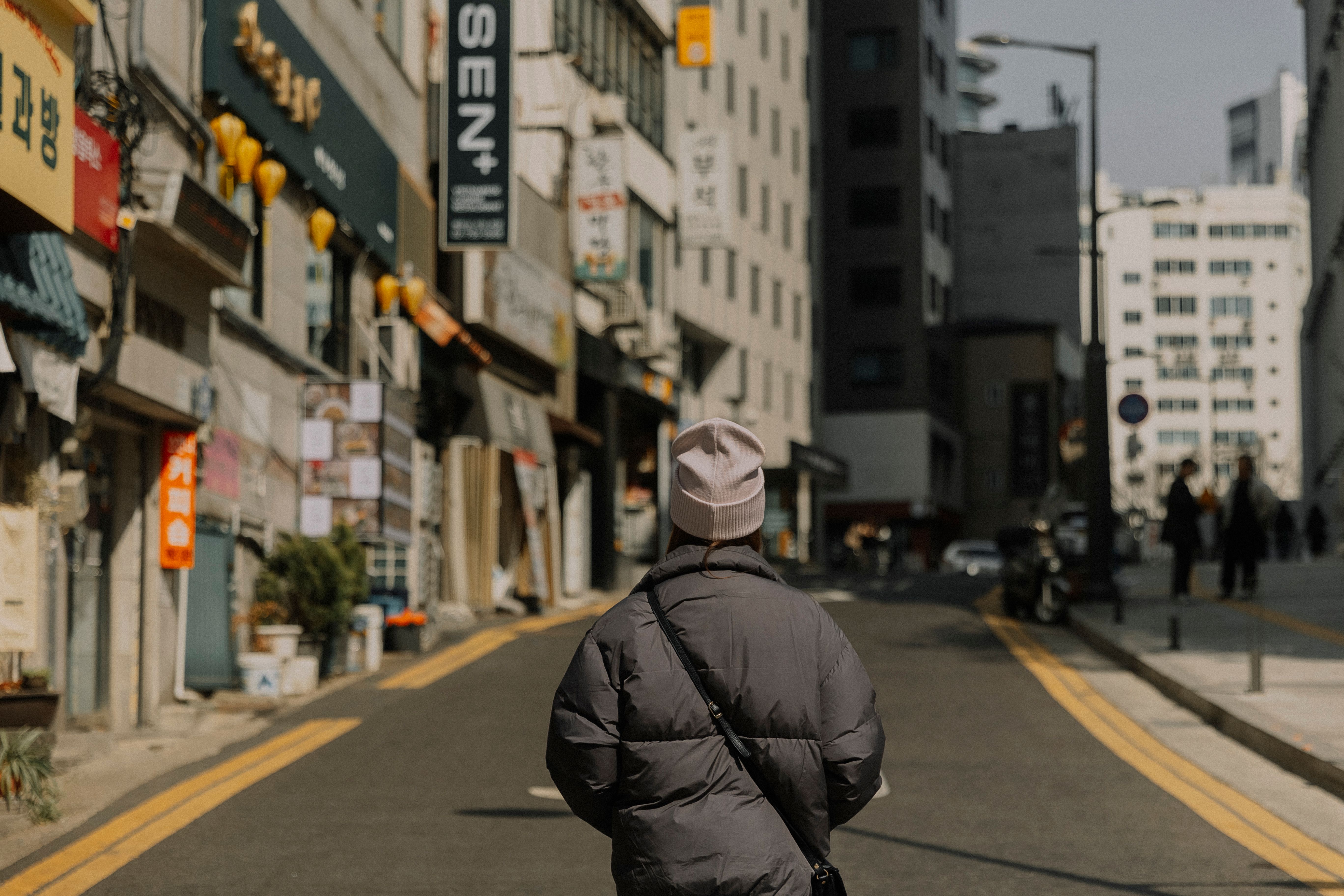 A woman in a winter jacket walks through a city's urban street, capturing modern architecture and shops.