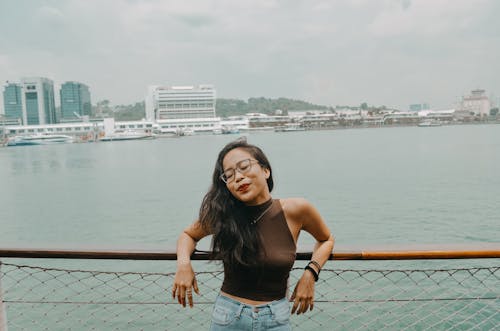 Free Photo of Woman With Her Eyes Closed Wearing Brown Top and Blue Denim Jeans Leaning Backwards on Wooden Railing with Harbor in the Background Stock Photo
