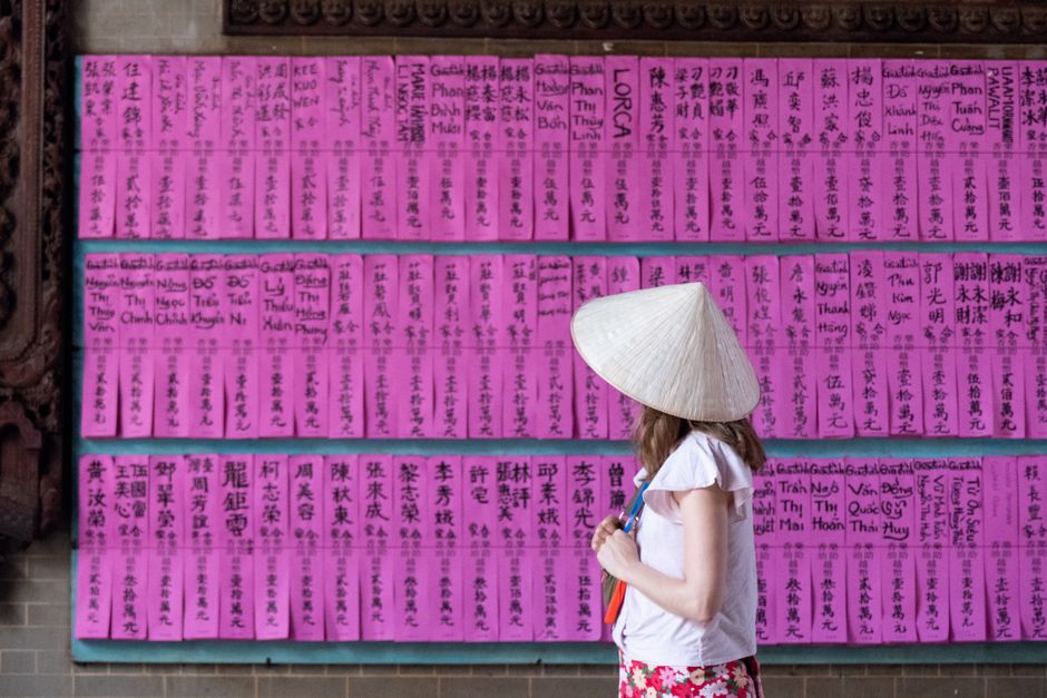 Woman in a conical hat observes pink donation notes at a Vietnamese temple.