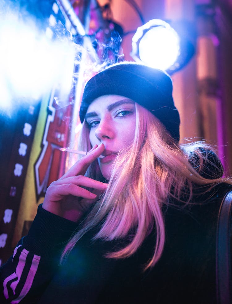 Portrait Photo Of Woman In Beanie Smoking A Cigarette While Under A Bright Light