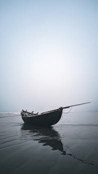 A solitary fishing boat rests on the calm sea shore under a foggy sky, creating a tranquil scene.