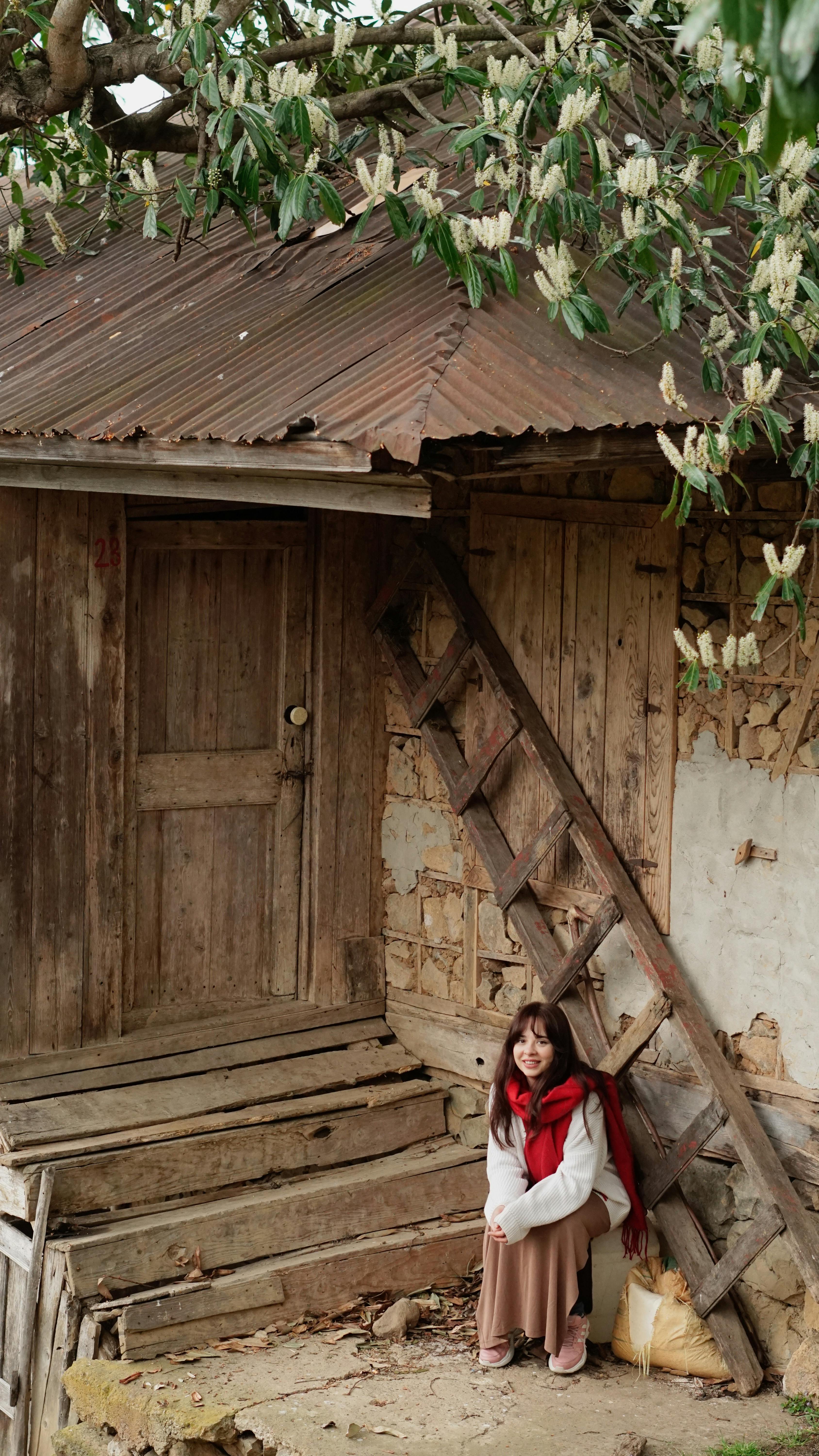 Young woman in red scarf sitting by rustic old house entrance with wooden ladder and greenery.