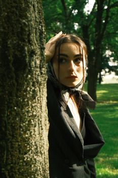 A fashion-forward brunette woman in a black suit jacket peers from behind a tree in a park setting.