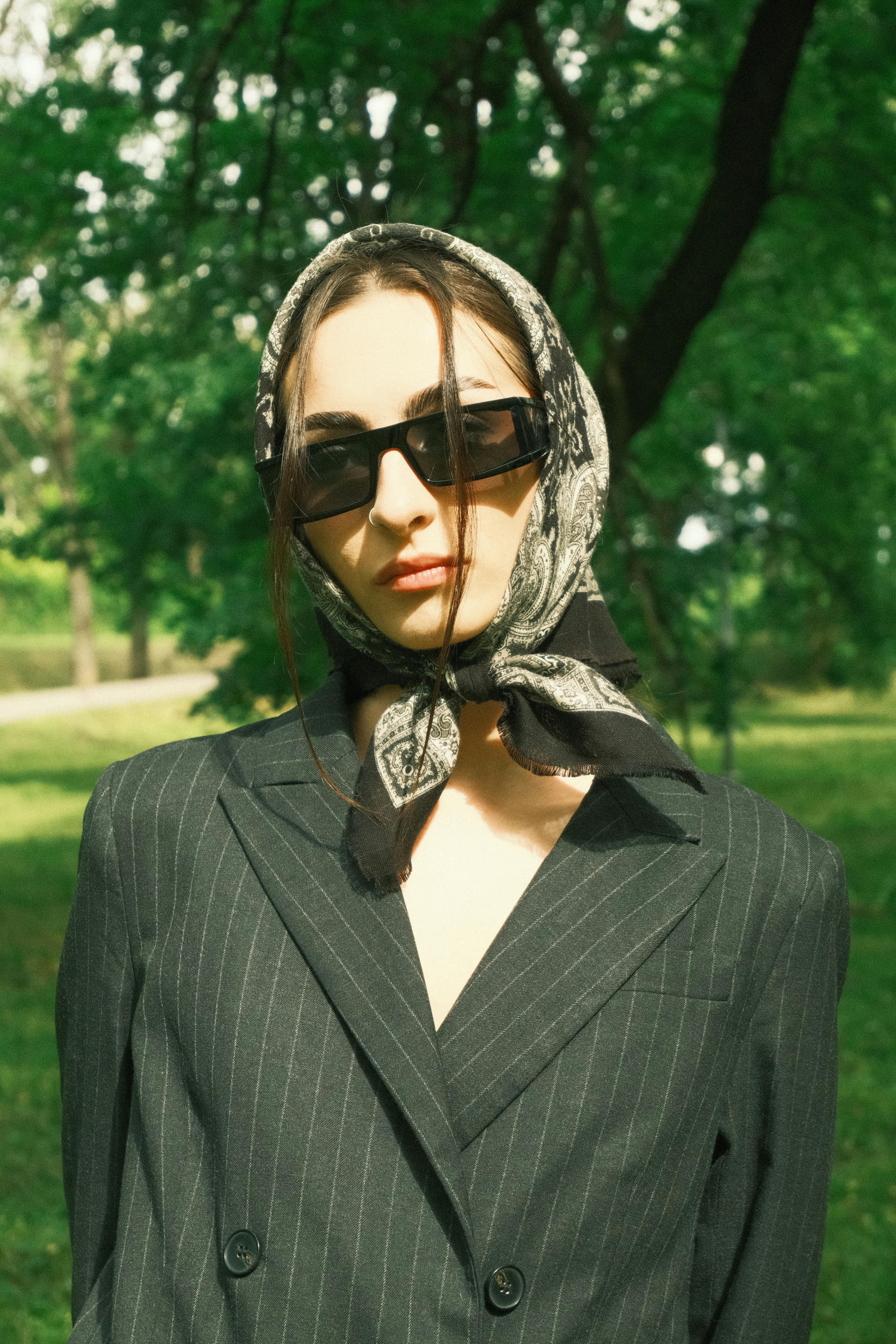 Stylish woman wearing sunglasses and a headscarf in a pinstripe jacket, set against a lush green background.