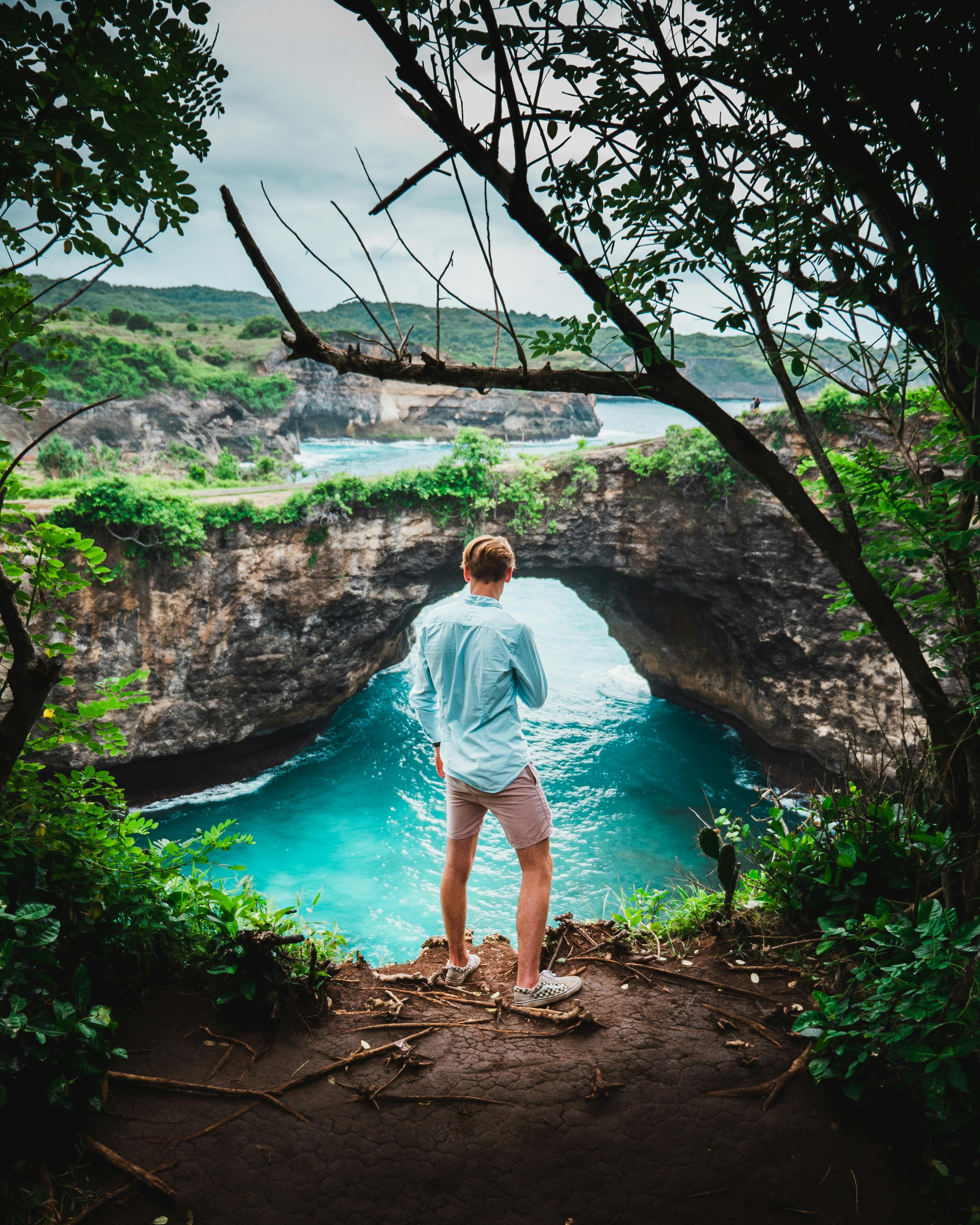 在印度尼西亚巴厘岛，一名男子站在悬崖上，欣赏着海拱门和碧绿的海水