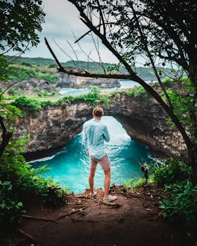在印度尼西亚巴厘岛，一名男子站在悬崖上，欣赏着海拱门和碧绿的海水