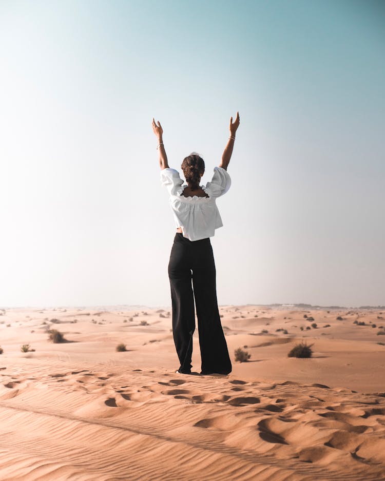 Back View Photo Of Woman With Her Hands Up Standing On Desert Sand