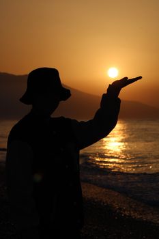 Silhouette of a person wearing a hat with raised hand against a golden sunset sky over the sea.