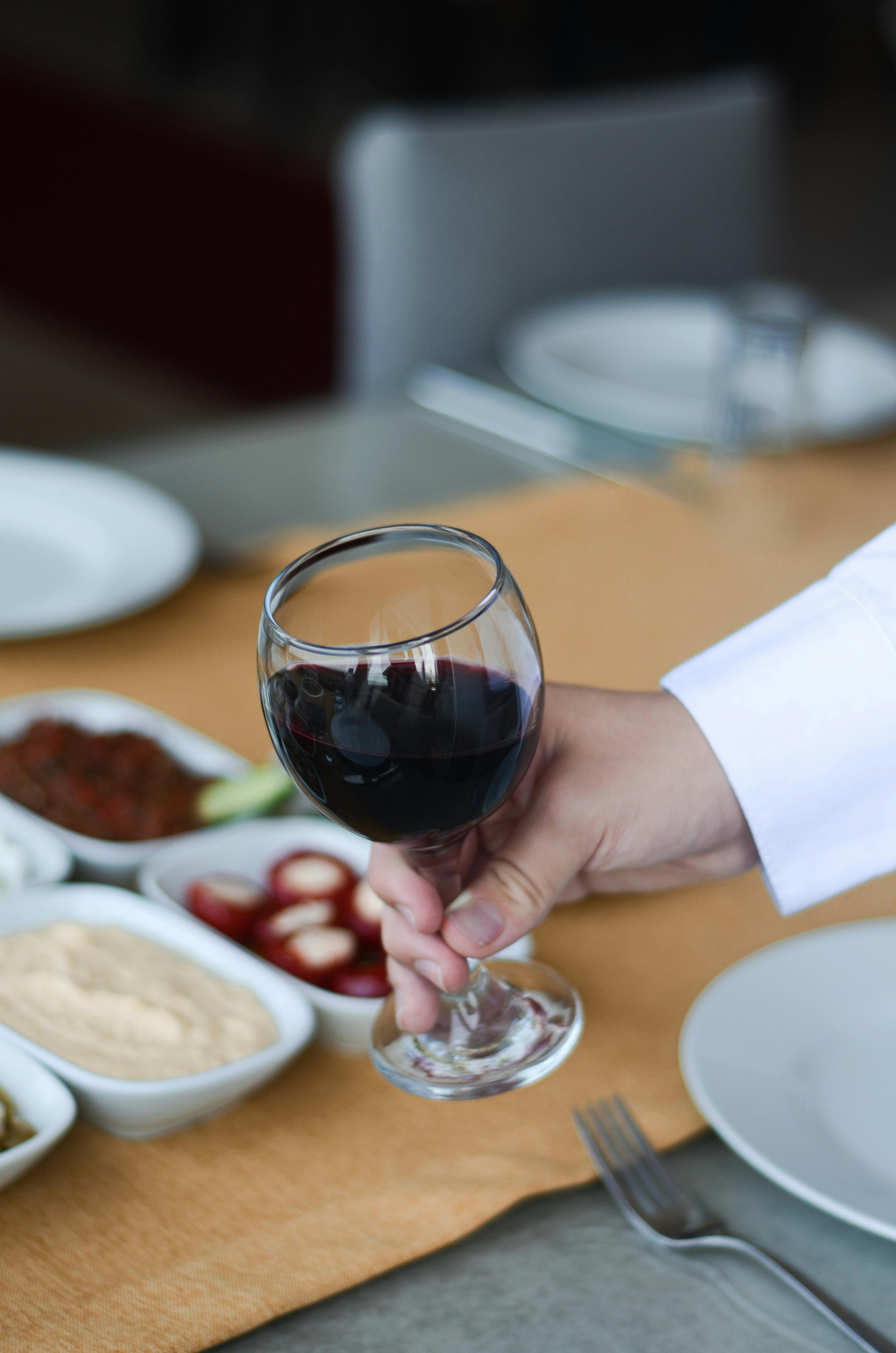 A close-up of a hand holding a glass of red wine at a dining table, ready for a meal.