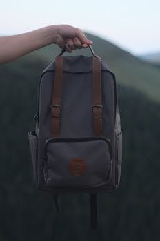 A person holding a stylish gray backpack with leather straps, set against a natural outdoor background.