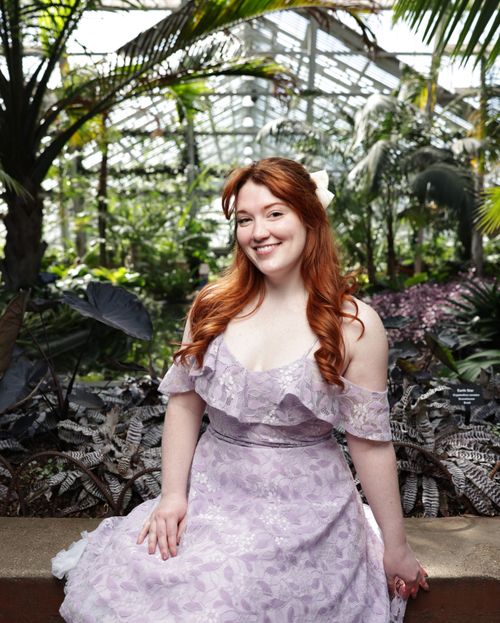 Free A young red-haired woman smiles brightly in a lush garden setting, wearing an elegant ivory dress. Stock Photo