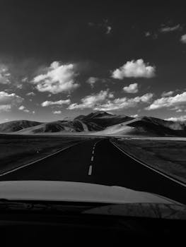 Driving down a deserted highway through mountainous terrain under a cloudy sky in black and white.