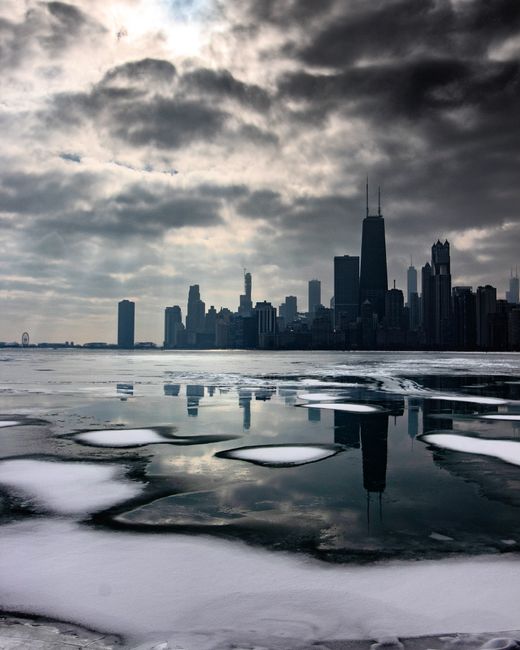 Stunning winter view of Chicago skyline reflected on ice-covered lake with dramatic cloudy sky.