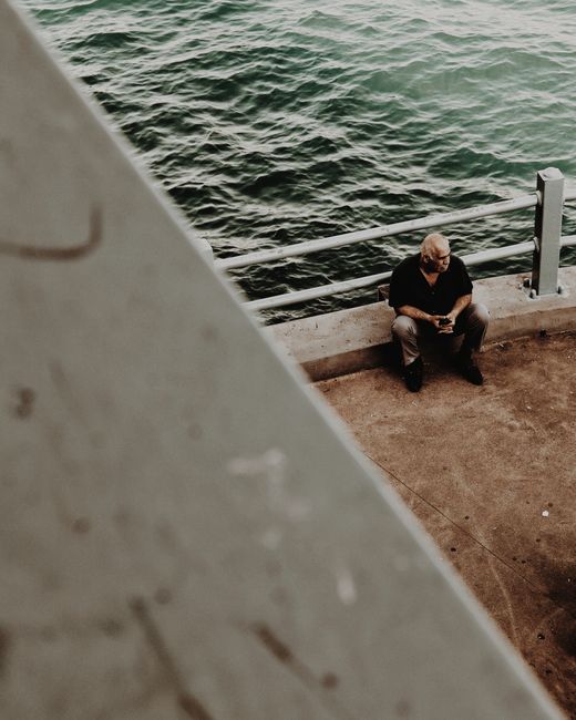 Elderly man sitting alone by a guard rail with water view, embodying solitude.