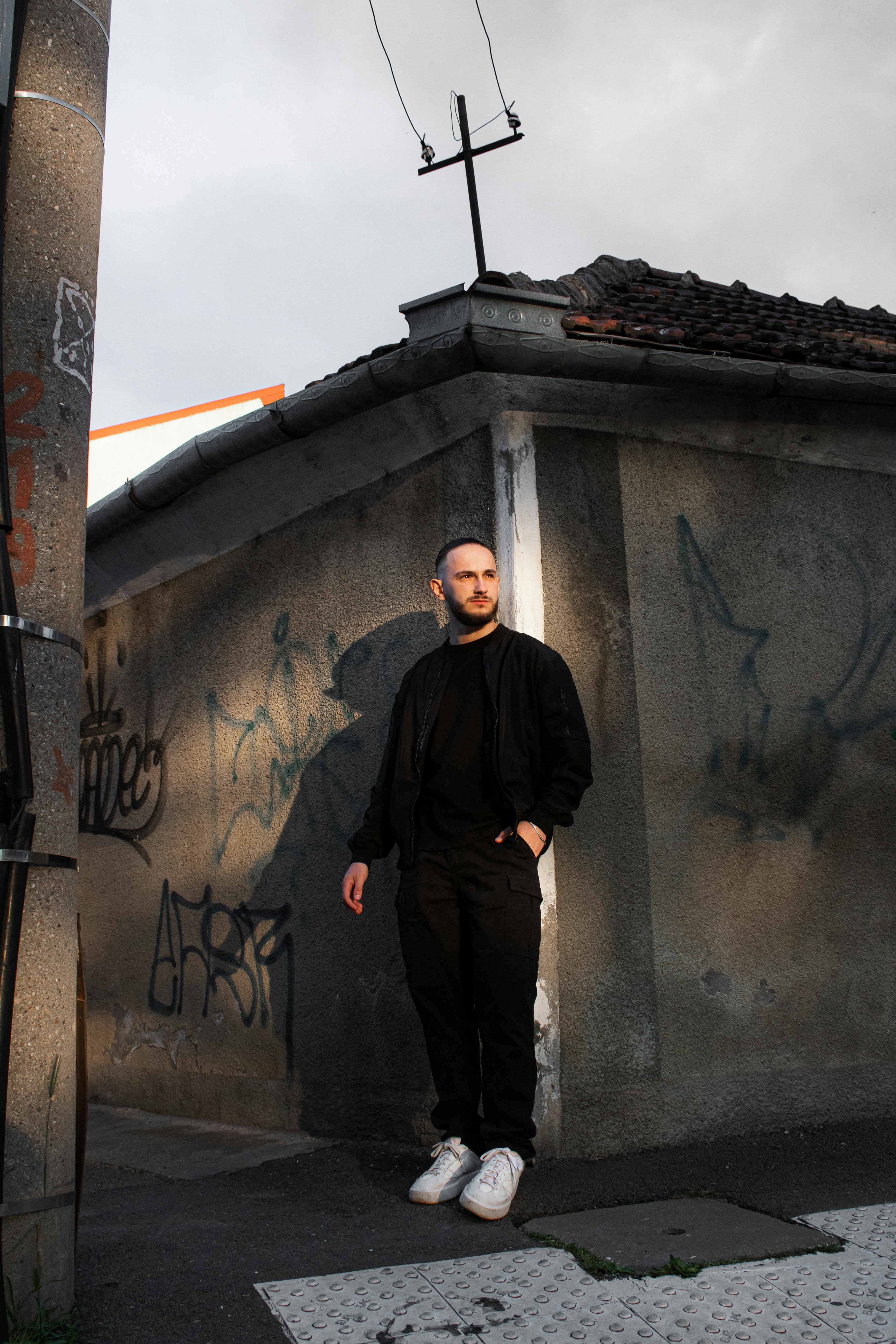 Stylish man in black jacket stands against graffiti wall in urban setting, exuding confidence.