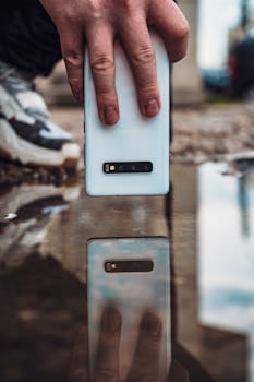 A hand holding a smartphone capturing its reflection in a puddle.