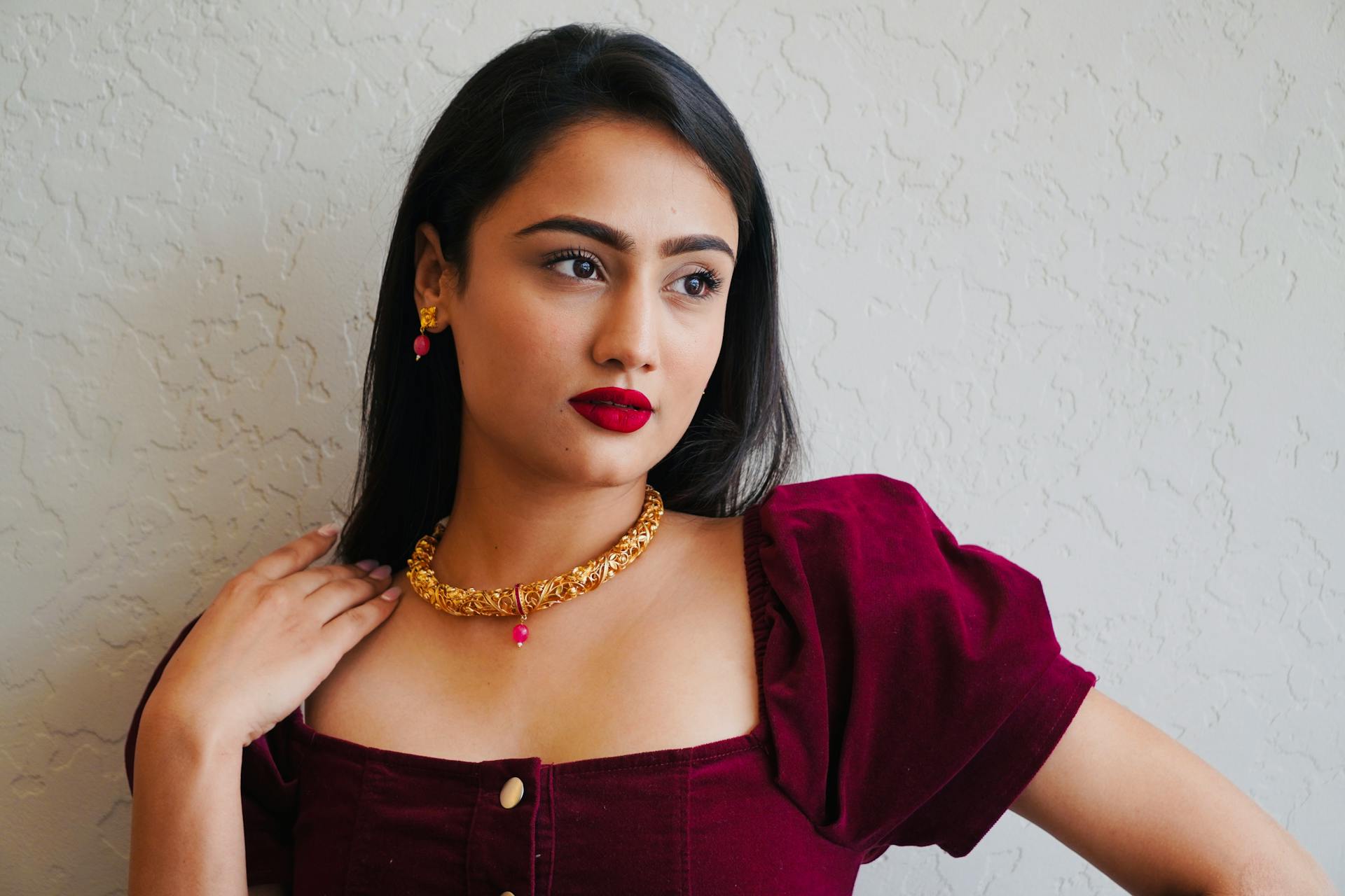 Stylish woman with red lipstick and gold jewelry posing elegantly indoors.