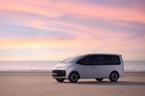 The sea under a pink sunset and the left front side view of the Hyundai Staria Hybrid.
