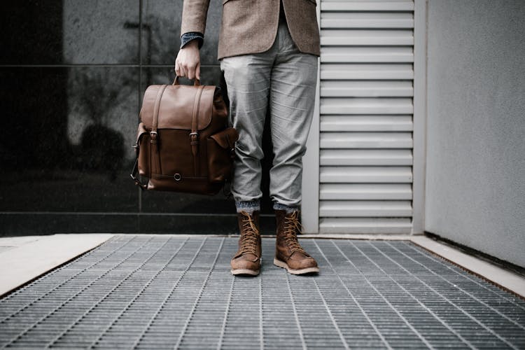 Photo Of Man In Brown Blazer,Gray Pants, And Brown Boots Holding Brown Leather Bag Standing Outside Building