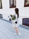一位身穿白色连衣裙的黑发女子，在阳光明媚的人行道上拿着一束色彩缤纷的花